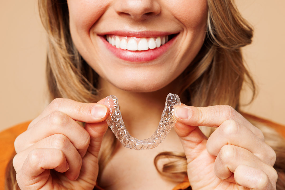 Close up cropped young woman wear orange shirt casual clothes hold in hand invisible transparent aligners, invisalign bracer smile isolated on plain pastel light beige background. Lifestyle concept
