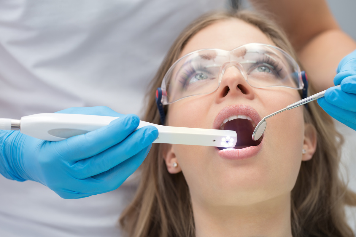 Attractive girl with opened mouth in protective eyewear. Next to her there is a dentist in a white uniform with blue latex gloves. He diagnoses her teeth with a LED intraoral camera and a stem mouth mirror. Horizontal.