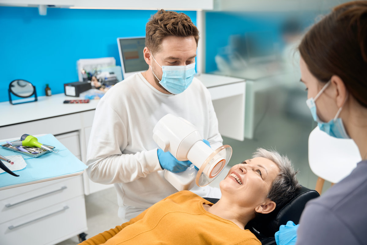 Aesthetic dentist in protective face mask holding portable dental x-ray near female patient, making image of teeth to check problem zones and find broken fillings or teeth with caries